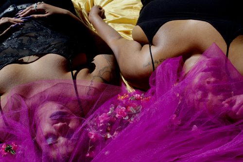 “Floral Refractions” –two Black womxn lay on gold satinwearing black clothing, they are posed with flowers neartheir faces, which are obscured bypurple tulle, their faces are adorned with indigo makeup.