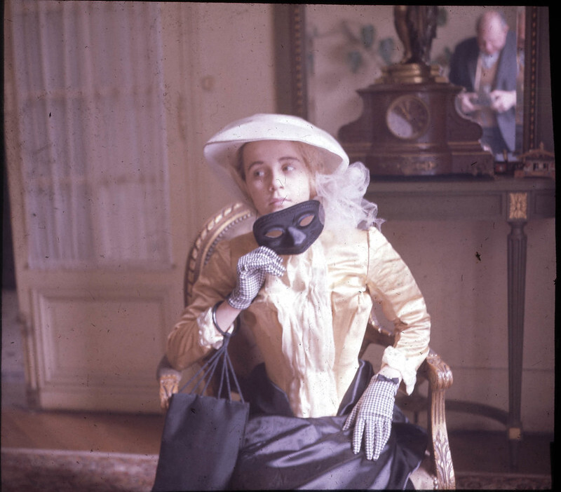A photograph of young woman in a yellow and black period dress holds a black mask.