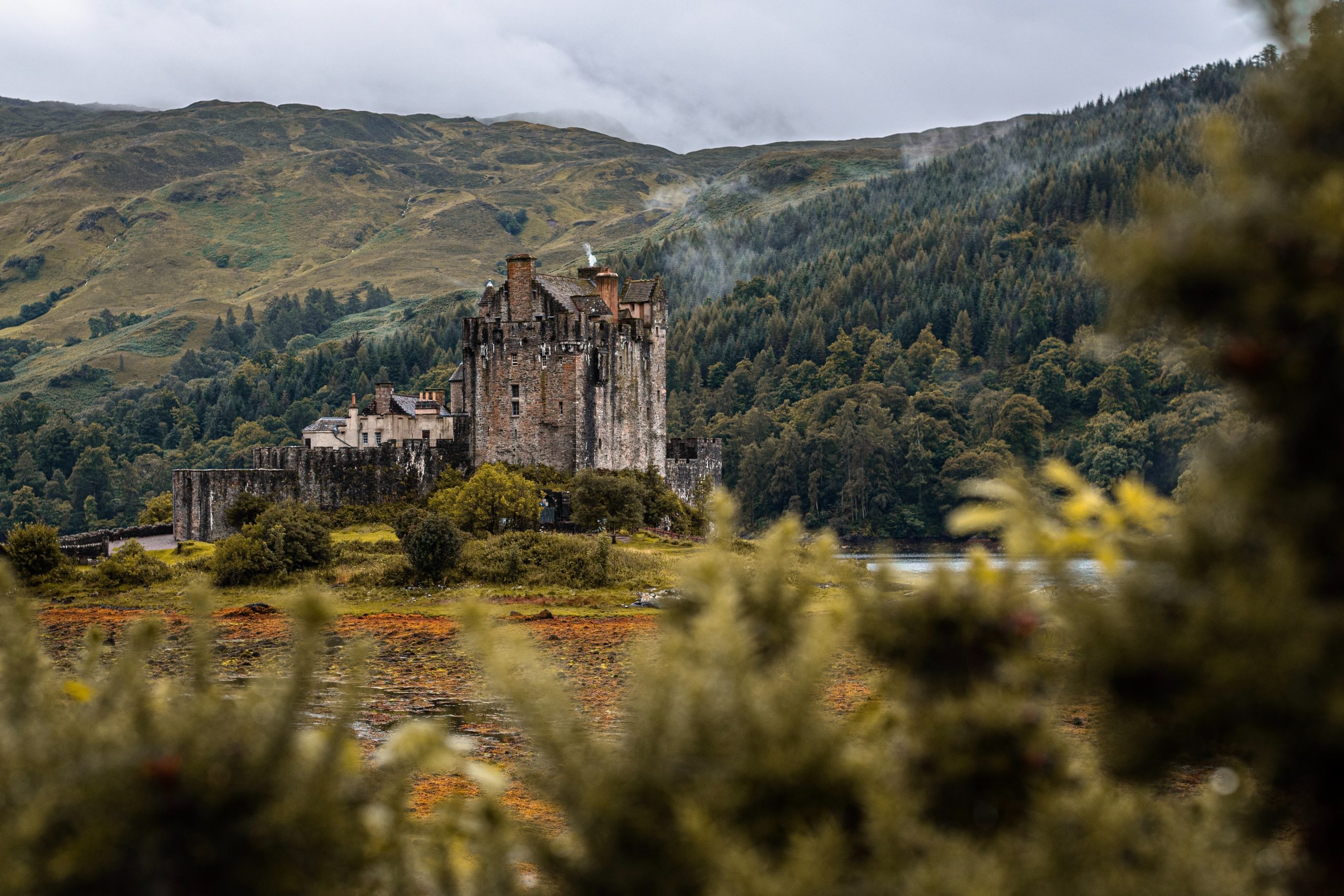 A castle in a clearing in front of the wooded hills of the Scottish highlands.